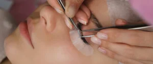 A lash glue bottle held at the tip with precision tweezers against a black background